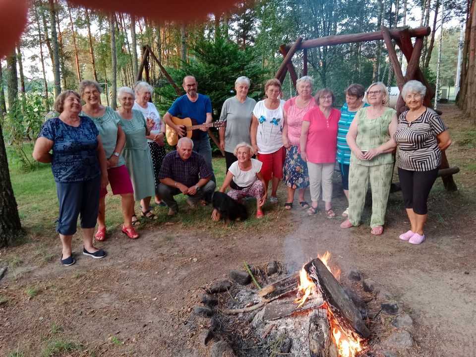 Seniorzy pozują do wspólnego zdjęcia przy ognisku, w tle drewniana huśtawka