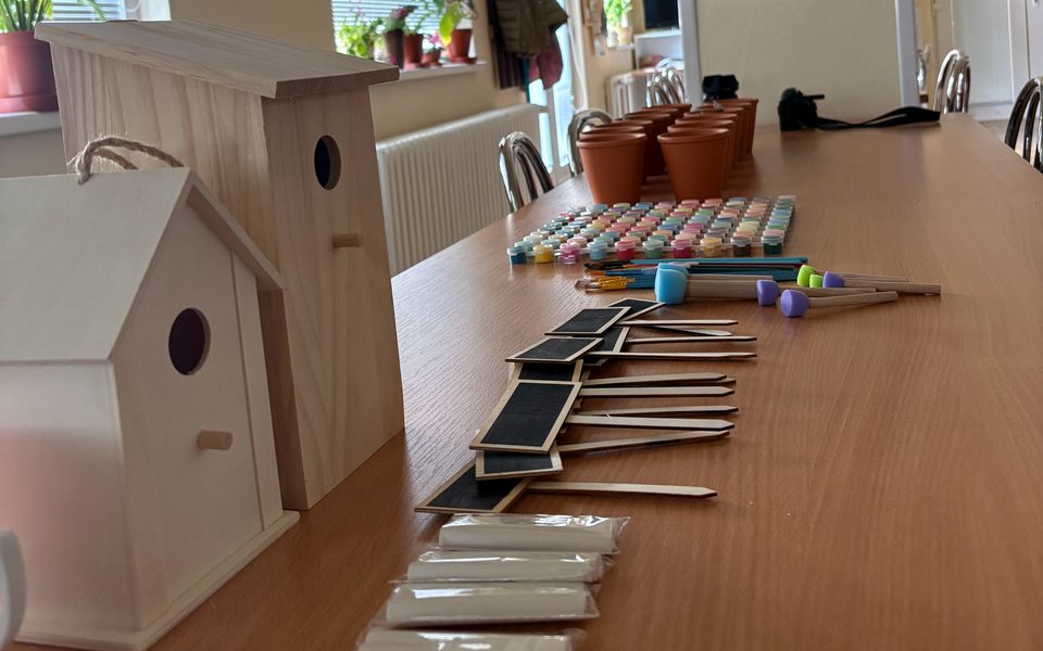 A long wooden table in a bright hall, set out with two unpainted wooden birdhouses, small paint pots, brushes, chalkboard plant labels, chalk and terracotta flower pots.
