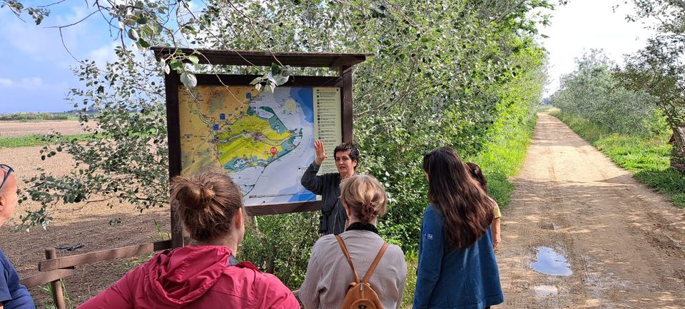 A woman points to a map of the Ebro Delta on an information board while the group listens; a sandy track leads away to the right.