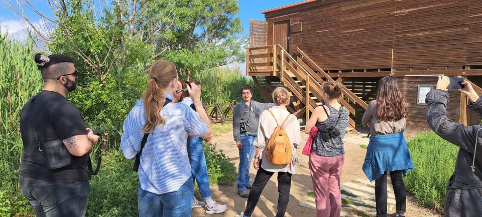 A woman with binoculars at her chest speaks to a group of visitors in front of a wooden observation building with an outside staircase.
