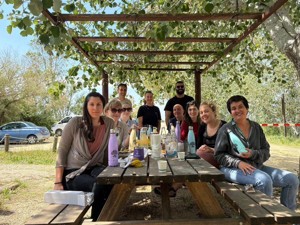 Project partners gathered around a wooden picnic table under a leafy pergola in the Ebro Delta, with ROOTED water bottles on the table.